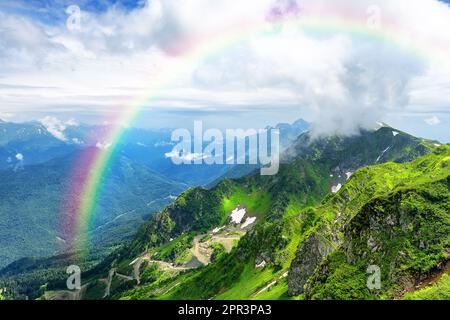 Amazing mountain landscape on sunny summer day with a rainbow in the sky. Mountains with green grass covered with snow against a cloudy sky. Stock Photo