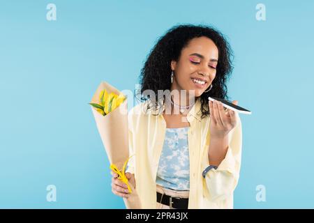 positive african american woman with yellow tulips sending voice message on smartphone isolated on blue,stock image Stock Photo