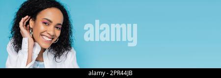 portrait of happy african american woman with perfect skin fixing wavy hair and smiling at camera isolated on blue, banner,stock image Stock Photo