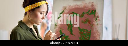 Focused young woman using her smartphone while creating abstract painting, standing in living room at home Stock Photo