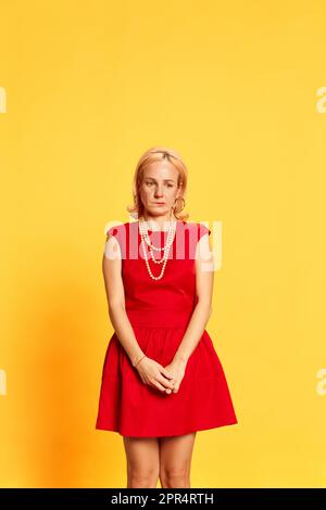 Portrait of blond woman in red dress looking down with a guilty face camera with shocked face on yellow background. Shyness, uncertainty Stock Photo