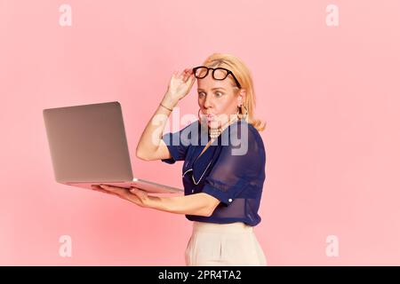 Portrait of beautiful blond young woman, in retro clothes holding laptop with shocked face over pink studio background. Business lady Stock Photo