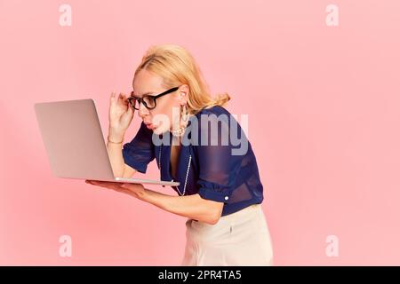 Portrait of harming young woman, in retro clothes and glasses holding laptop with shocked face over pink background. Business progress Stock Photo