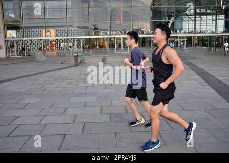 Two male joggers run past shopping mall The Shoppes At Marina Bay Sands by Marina Bay, Singapore Stock Photo