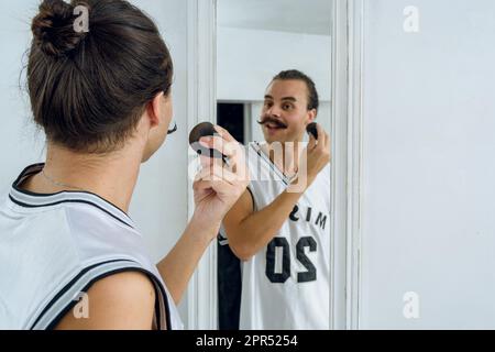happy cheerful young gender non-binary person doing makeup in her room, standing in front of the mirror with a sponge, talking and laughing, copy spac Stock Photo