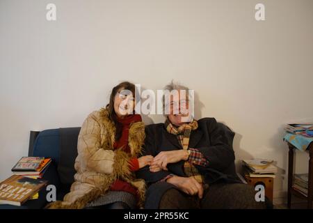 Smiling, happy and positive retired couple, in love, sitting on sofa, holding hands -  wearing winter clothes, coat and scarf, keeping warm; indoors Stock Photo