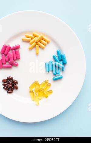 Food supplements of bright color lie on a white plate on a blue background. Concept of health support and lack of vitamins. Vertical photo, close-up, Stock Photo