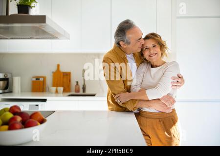 Happy loving senior spouses in casual outfits embracing, enjoying time together at cozy kitchen, man kissing his wife Stock Photo