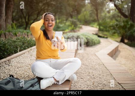 Wow. Excited young brazilian lady reading great news on smartphone and touching head, sitting in park outdoors Stock Photo