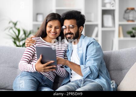 Happy loving indian couple sitting on couch, using digital pad Stock Photo