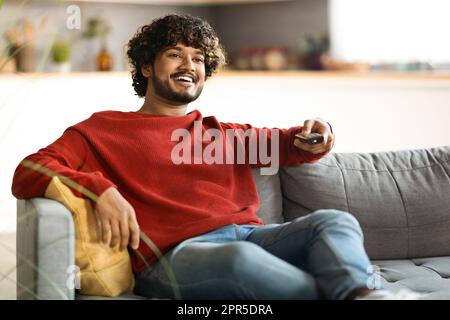 Weekend Pastime. Cheerful Indian Guy Watching Tv At Home, Holding Remote Controller Stock Photo