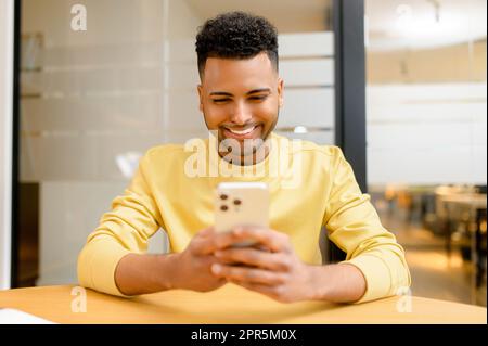Cheerful young Indian guy spends leisure scrolling news feed on the smartphone, handsome guy holds mobile phone, looks at the screen, websurfing, shopping, chatting in the social networks Stock Photo