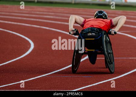 rear view male athlete in racing wheelchair riding on red track stadium, summer para athletics championships Stock Photo