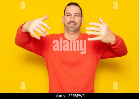 Smiling bearded hispanic man 40s wearing casual pullover posing stretching hands for hug while noticing someone looking at camera isolated on yellow Stock Photo