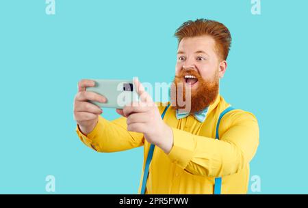 Cheerful fat man on light blue background makes selfie shot on front camera of mobile phone. Stock Photo
