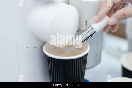 Woman making a cup of hot coffee with capsule coffee machine. Woman hand holding frothed milk dispenser of capsule coffee machine on table. Espresso coffee maker. Morning drink. Modern home equipment. Stock Photo
