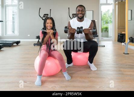 Young short curly black hair man and his daughter lifting barbell with both hands while sitting on yoga ball. Happy family enjoy holiday together in fitness center. Stock Photo