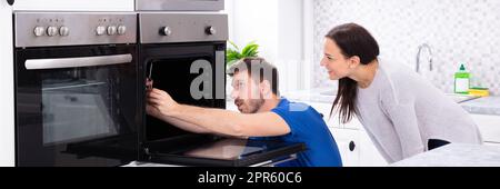 Technician In Overall Fixing Oven In Kitchen Stock Photo