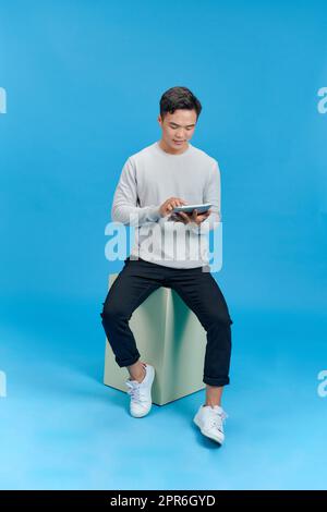 Happy young man sitting on geometry cube and using a tablet over blue background Stock Photo