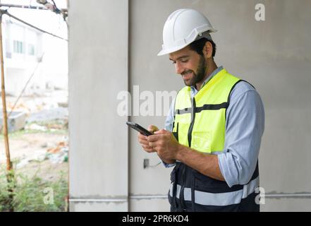Young engineer in hardhat and safety vest typing text massage on mobile phone. Work environment of engineers at the construction site. Stock Photo
