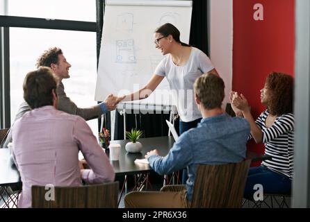 Great presentation. Shot of a two people shaking hands in a boardroom while colleagues look on. Stock Photo