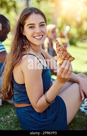 Its a slice of happiness. Portrait of a young woman eating pizza while out on a picnic with friends. Stock Photo