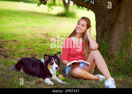 Enjoying a carefree day at the park. Shot of a young woman reading a book while sitting with her dog under a tree. Stock Photo