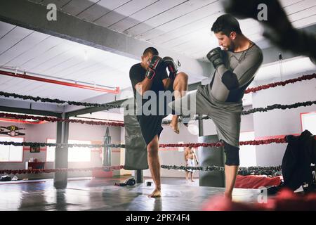 Using some kicks to mix it up. Shot of two young male boxers facing each other in a training sparing match inside of a boxing ring at a gym during the day. Stock Photo