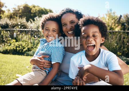 Excited little boy with afro sitting outside on the grass with his mother and brother. Energetic african american family spending time outdoors at the park or their backyard. Mom embracing two sons Stock Photo