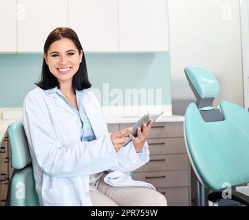 A beautiful, confident female dentist sitting in her doctors rooms. A confident, brunette woman working on a digital tablet while sitting in her office. Dental health resources are easy to find online Stock Photo
