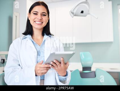 Young female caucasian dentist wearing a labcoat and smiling while using a digital tablet in her office. Dental hygiene is important to your wellbeing Stock Photo
