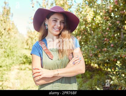 A beautiful female farm worker standing with her arms crossed on a fruit farm during harvest season. Young happy farmer between fruit trees in summer. Agricultural industry growing produce Stock Photo