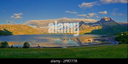 Countryside landscape in Nordland, Bodo, Norway with.glistening waters on a lake near a majestic mountainscape during sunset. Blue sky with lush green grass and a little house on a river bank Stock Photo