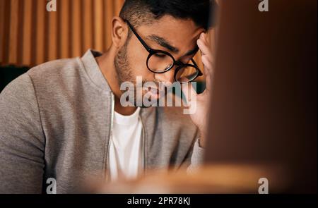 Young stressed indian man or student struggling with headache while browsing on a laptop in a cafe. One tired and frustrated guy suffering with burnout and pressure while studying or working freelance Stock Photo