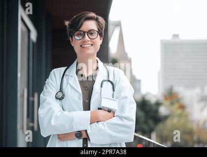 A doctor thats here to help. a young female doctor standing with her arms crossed against a city background. Stock Photo