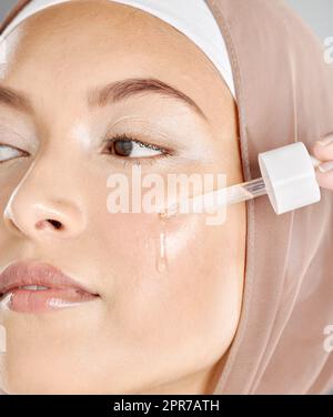 A young woman applying antiaging facial serum to her face and skin. Beautiful muslim girl wearing a hijab trying retinol with a dropper as her skincare routine and beauty treatment regime Stock Photo
