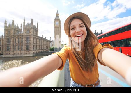 Smiling tourist girl taking self portrait in London, UK. Selfie photo of happy woman traveling in London with Big Ben tower, Westminster palace and double decker red bus on summer sunny day. Stock Photo