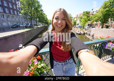 Pretty girl in Netherlands on sunny day. Smiling young woman takes selfie picture on canal in The Hague, Netherlands. Stock Photo