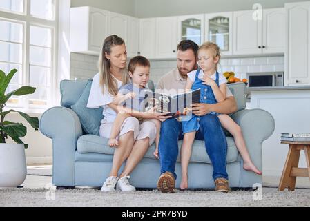Caucasian family reading a book together on the couch at home. Mother and father teaching their little son and daughter how to read. Brother and sister learning their alphabet with their parents Stock Photo