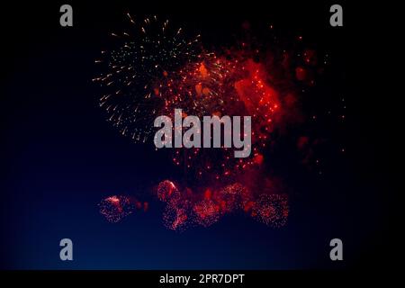 Red and orange fireworks with green sparks inside and smoke, below you can see small fireworks against the night sky Stock Photo