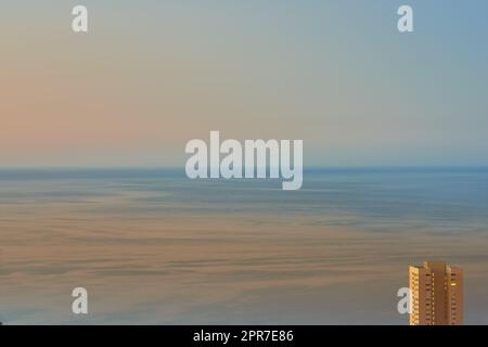 Calm waters during sunset with a hotel building overlooking the ocean. Aerial view of the sea during dusk. Copy space of small waves on calm water in the sea, bright afterglow in a clear summer sky Stock Photo