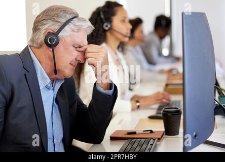 The solution is nowhere in sight. a mature call centre agent looking stressed out while working in an office with his colleagues in the background. Stock Photo