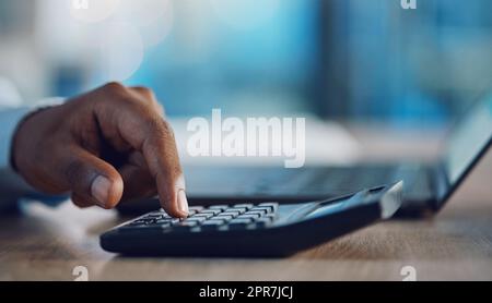Closeup hands of african man using a calculator while sitting at his desk. African american business man calculating figures while working late at night in his office. Putting in overtime after hours Stock Photo