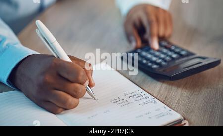 Closeup hands of african man writing in his diary while using a calculator. African american business man calculating figures while working late at night in his office. Putting in overtime after hours Stock Photo