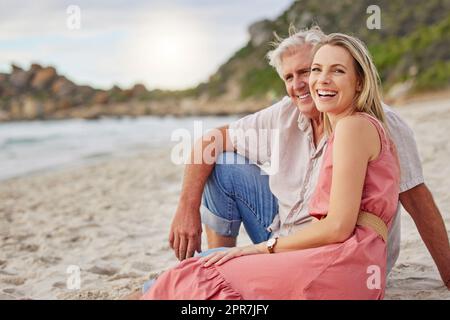Portrait of a senior caucasian man smiling and spending time with his daughter on vacation at the beach while sitting on the sand Stock Photo