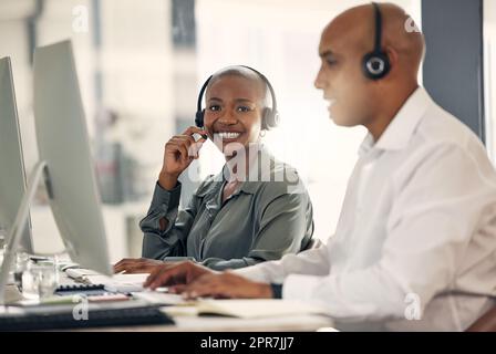 Portrait of happy young african american call centre telemarketing agent talking on a headset while working on a computer in an office alongside a colleague. Confident friendly female consultant operating helpdesk for customer service and sales support Stock Photo