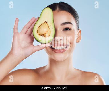 Beautiful young mixed race woman with an avocado isolated in studio against a blue background. Her skincare regime keeps her fresh. For glowing skin, eat healthy. Packed with vitamins and nutrients Stock Photo