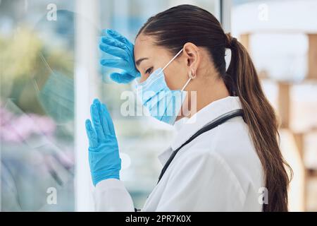 Young stressed and overworked doctor wearing mask and gloves while standing at a window in a hospital or clinic. One female only looking worried, hopeless and anxious while struggling with a challenge Stock Photo