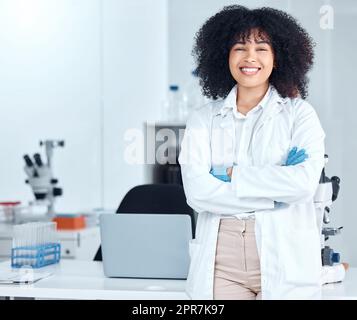 Portrait of a beautiful young african american woman with an afro wearing a labcoat and gloves while standing with her arms crossed in the laboratory. A mixed race female scientist smiling happily Stock Photo