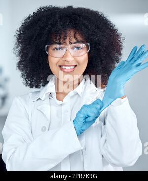 Portrait of young african american female scientist with afro hair wearing a labcoat and goggles while putting on gloves in the laboratory. A mixed race female scientist getting ready to conduct an experiment Stock Photo